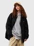 A woman with curly red hair wearing beige cap, black quilted jacket over gray sweatshirt and white collar, posing against white background