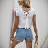 Rear view of a woman in a white embroidered top with ruffled sleeves and a bow-tie back, paired with distressed denim shorts, standing in an alley.