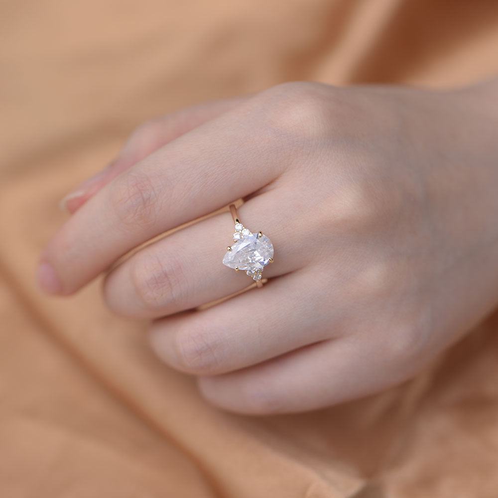 A woman wearing a delicate gold engagement ring with pear-cut crystal and accent diamonds, resting her hand against soft beige fabric