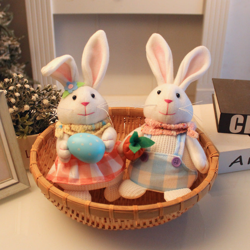 Two fabric bunny figurines in a wicker basket, one in a blue plaid outfit with a carrot, the other in a red-checkered dress holding an Easter egg.