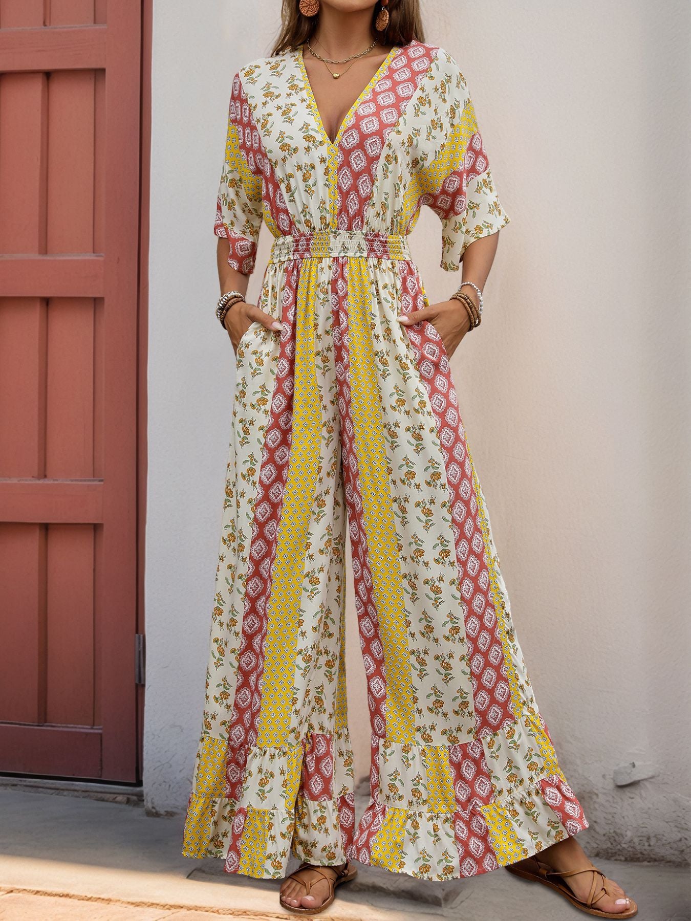 Woman in a patchwork floral jumpsuit with a cinched waist and pockets, wearing gold accessories and brown sandals.