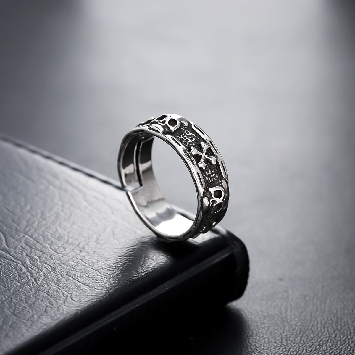 A close-up of a silver skull ring placed on a black leather surface, showcasing its polished finish and detailed cutout design.

