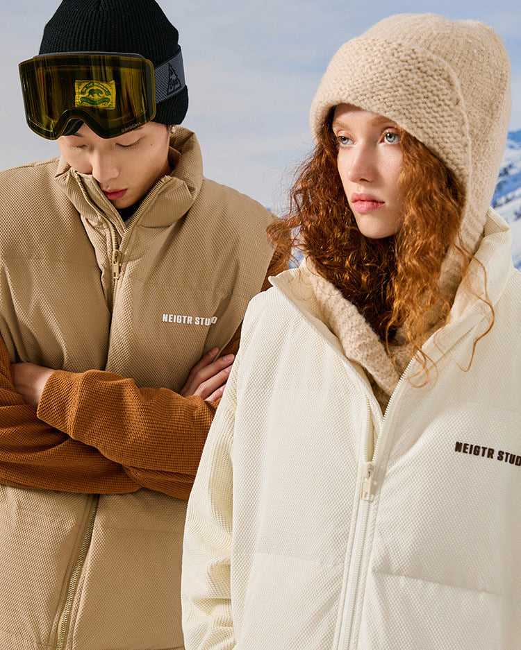Models wearing NEIGTR STUDIO outerwear - man in tan jacket with black beanie and woman in cream jacket with knit beanie against snowy backdrop