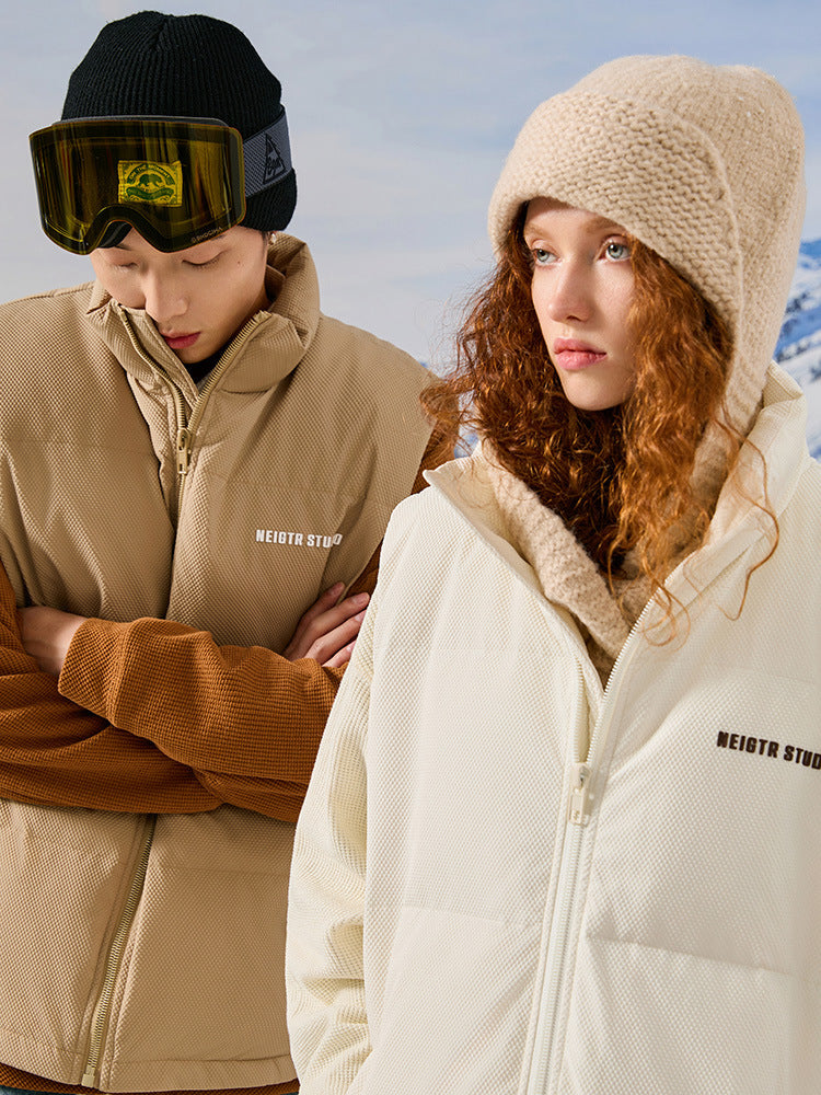 Models wearing NEIGTR STUDIO outerwear - man in tan jacket with black beanie and woman in cream jacket with knit beanie against snowy backdrop