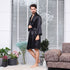 Side view of man in black silk robe standing barefoot against white blinds, with decorative plants and tweed sofa visible in the styled home setting