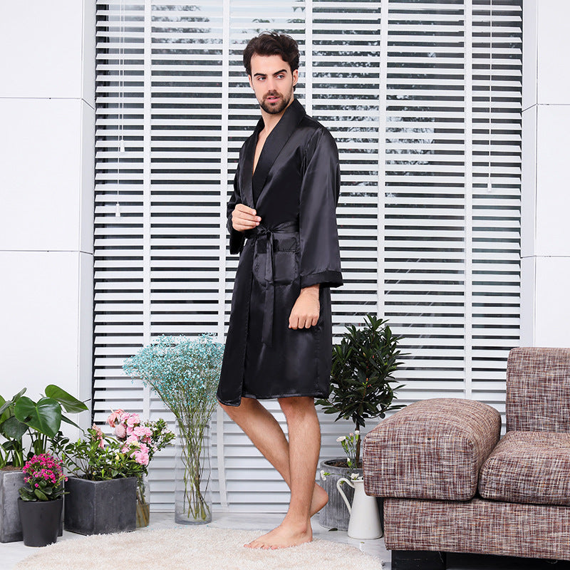 Side view of man in black silk robe standing barefoot against white blinds, with decorative plants and tweed sofa visible in the styled home setting
