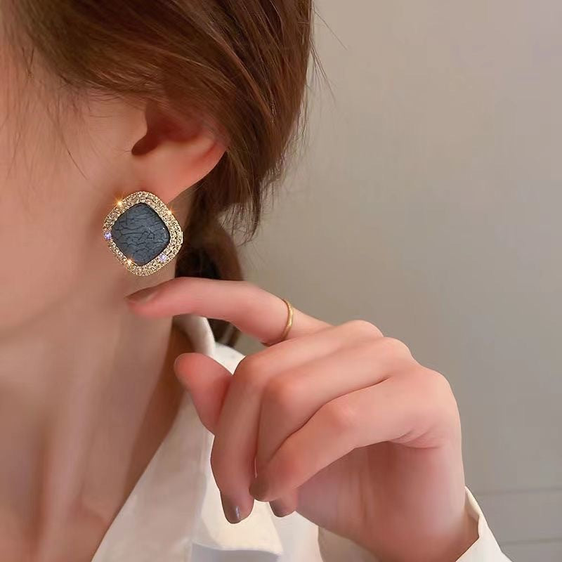 Square stud earring with blue crackle stone and crystal rim worn on ear, shown with delicate gold ring