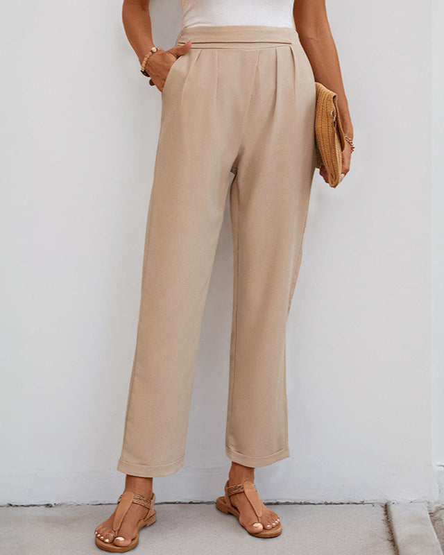 A woman dressed in beige straight-leg trousers with a fitted high waist, accessorized with a woven clutch and tan sandals.