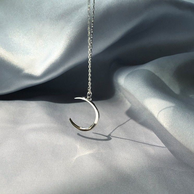 Silver crescent moon pendant on chain, photographed against draped silk fabric with shadow detail