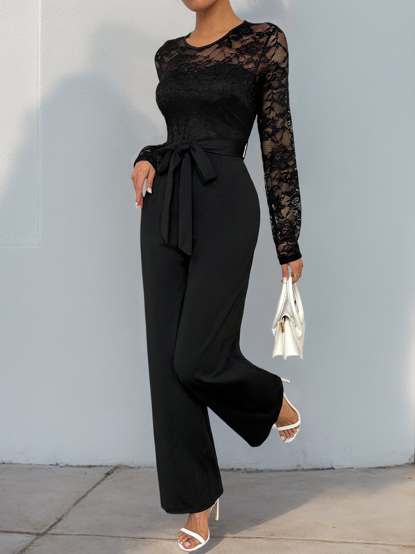 A side view of a woman in a black lace top jumpsuit with a cinched waist, holding a white handbag while stepping forward.