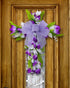 A purple and white floral cross wreath with a large bow, elegantly hanging on a rustic wooden door with intricate detailing.