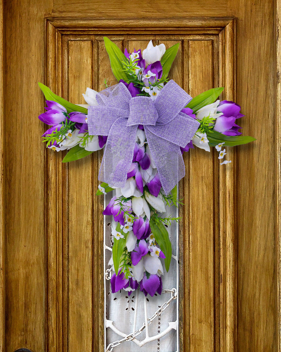 A purple and white floral cross wreath with a large bow, elegantly hanging on a rustic wooden door with intricate detailing.