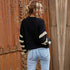 A woman with wavy blonde hair shown from behind wearing a black knit cardigan with cream striped sleeves and light blue jeans beside ornate wooden doors