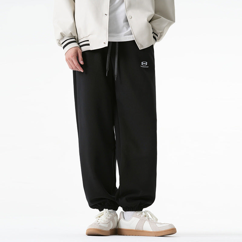 A model is wearing black jogger-style sweatpants with an elastic drawstring waist and cuffed ankles, paired with a cream varsity jacket and white sneakers
