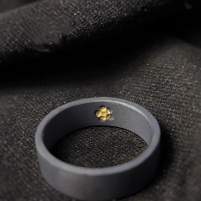 A single black ring with a small embedded gold accent inside, placed on textured black fabric.