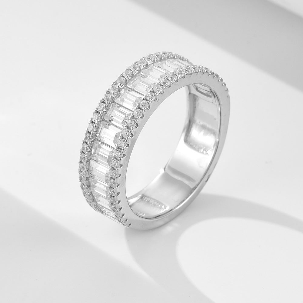 A silver eternity band with baguette-cut diamonds bordered by rows of round pavé diamonds, displayed on white surface in natural light