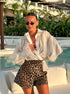 Woman with sunglasses near a pool, styling a sheer white blouse and leopard print shorts.
