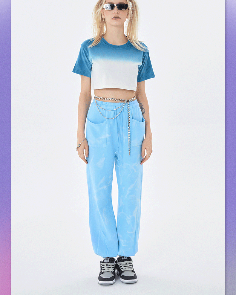 Blue tie-dye crop top with white panel, matching blue jogger pants, and gray sneakers; model wears white rectangular sunglasses