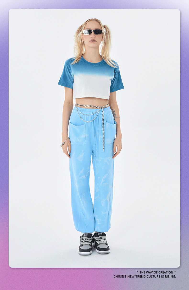 Blue tie-dye crop top with white panel, matching blue jogger pants, and gray sneakers; model wears white rectangular sunglasses