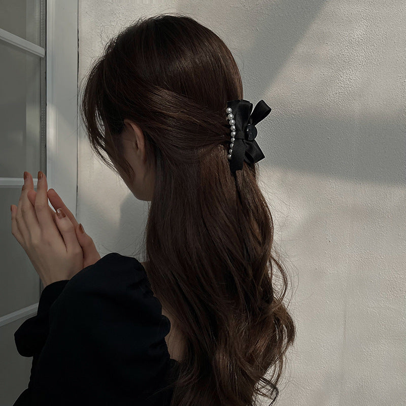 Half-up hairstyle with wavy brown hair, adorned with a black bow hair clip featuring pearl embellishments, against a white textured wall background