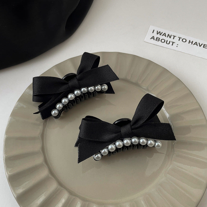 Two elegant black hair clips with pearl-studded centers and black satin bows, displayed on a textured cream-colored plate with a minimalist label