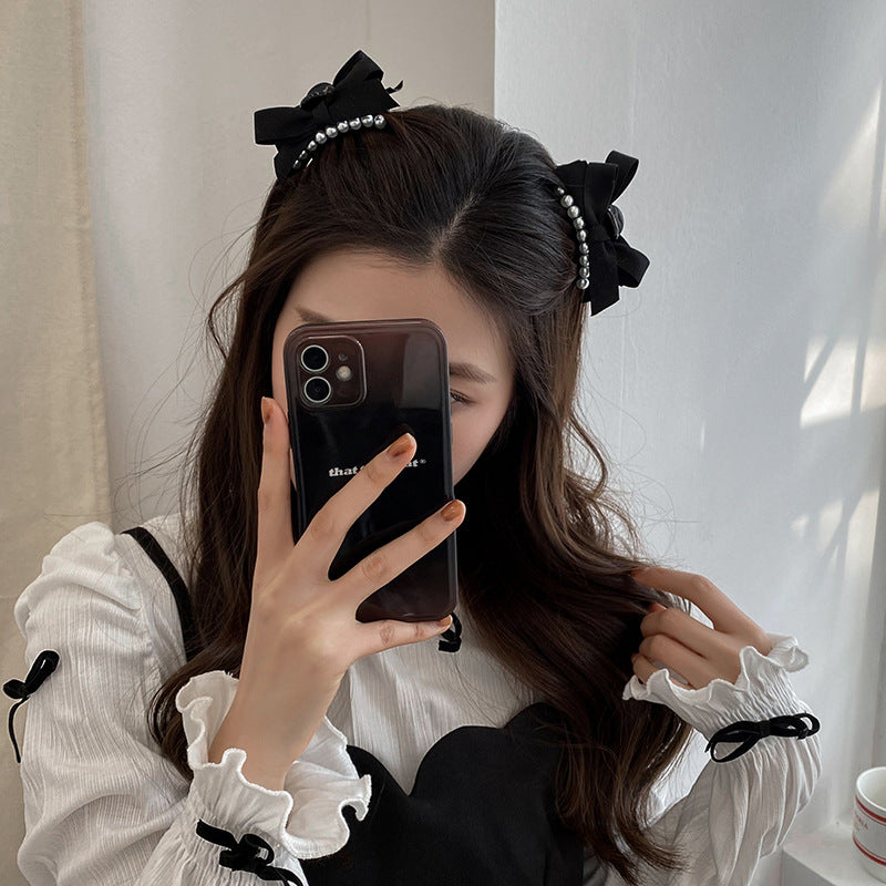 Mirror selfie of a woman wearing a white top, with black pearl bow hair clips holding back her brown hair, partially obscured by a black smartphone