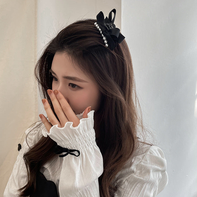 Side profile of a woman in a white top, wearing a black pearl bow hair clip, with her hand covering part of her face