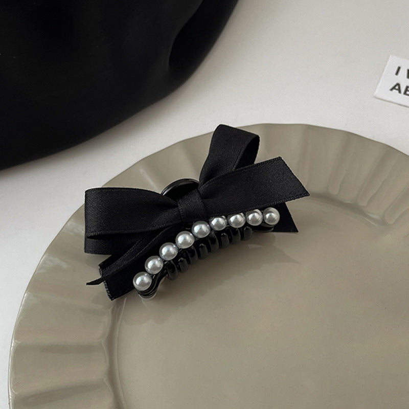Black pearl bow hair clip positioned on a scalloped-edge ceramic plate, with a partial view of a black object and a white label in the background