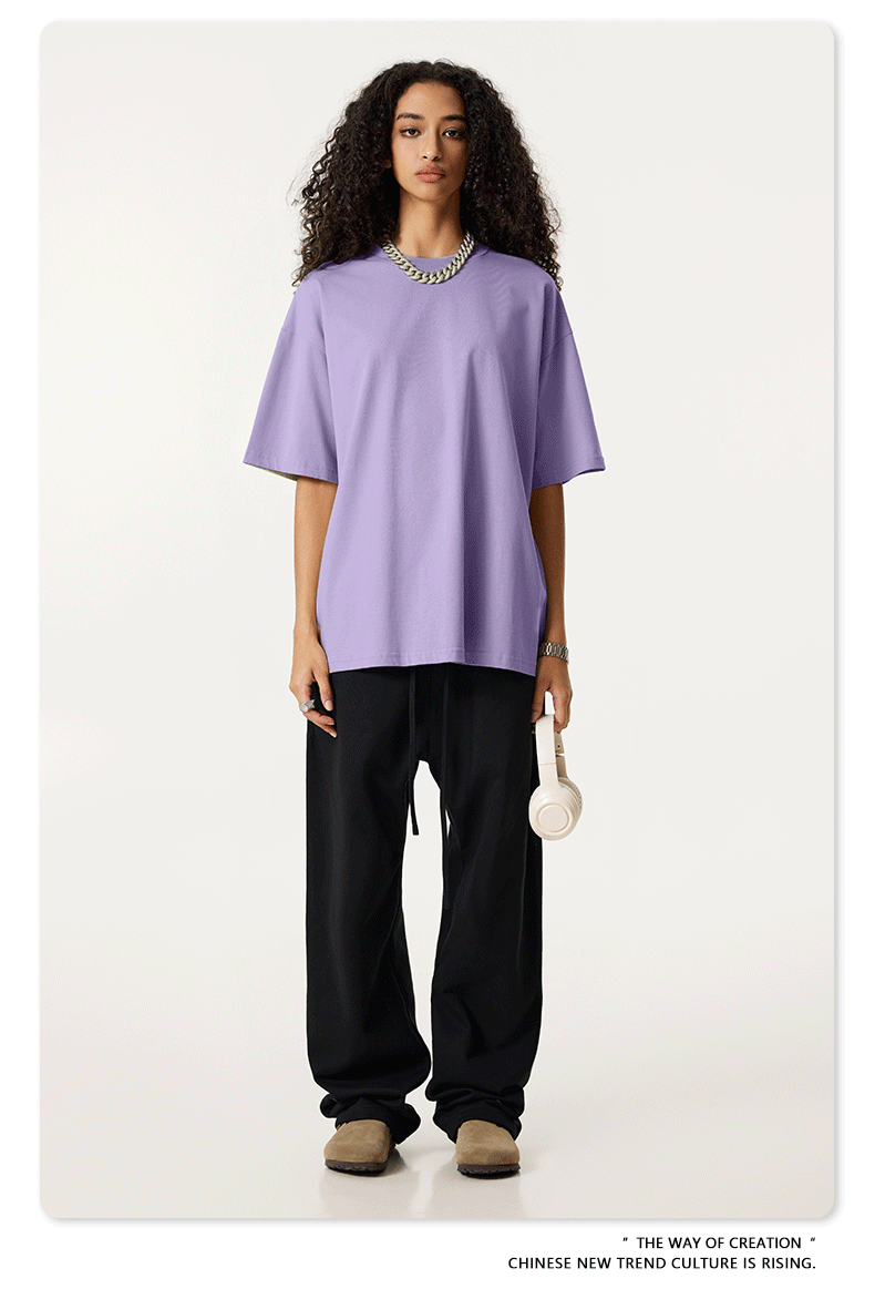 Female model in lavender oversized t-shirt paired with black pants, holding white headphones, wearing silver chain necklace and beige shoes