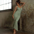 A woman wearing a sage green ruched mermaid dress with cutout detail poses against textured concrete wall in industrial setting