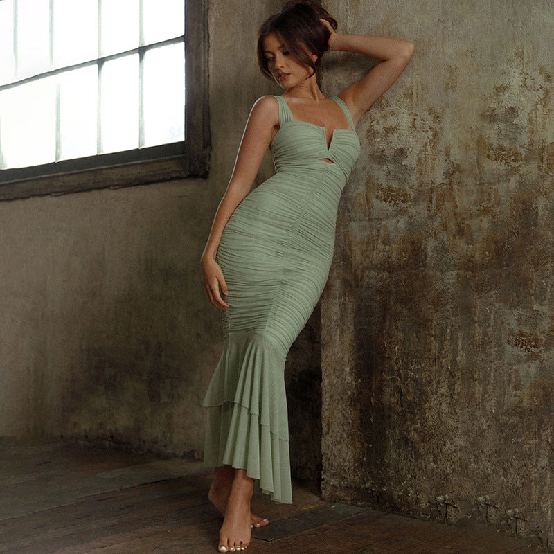 A woman wearing a sage green ruched mermaid dress with cutout detail poses against textured concrete wall in industrial setting