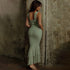 A woman showing back view of sage green ruched bodycon dress with tiered hem against concrete wall backdrop