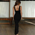 A woman in black fitted dress with wide straps shown from behind walking across wooden floor with white curtain backdrop