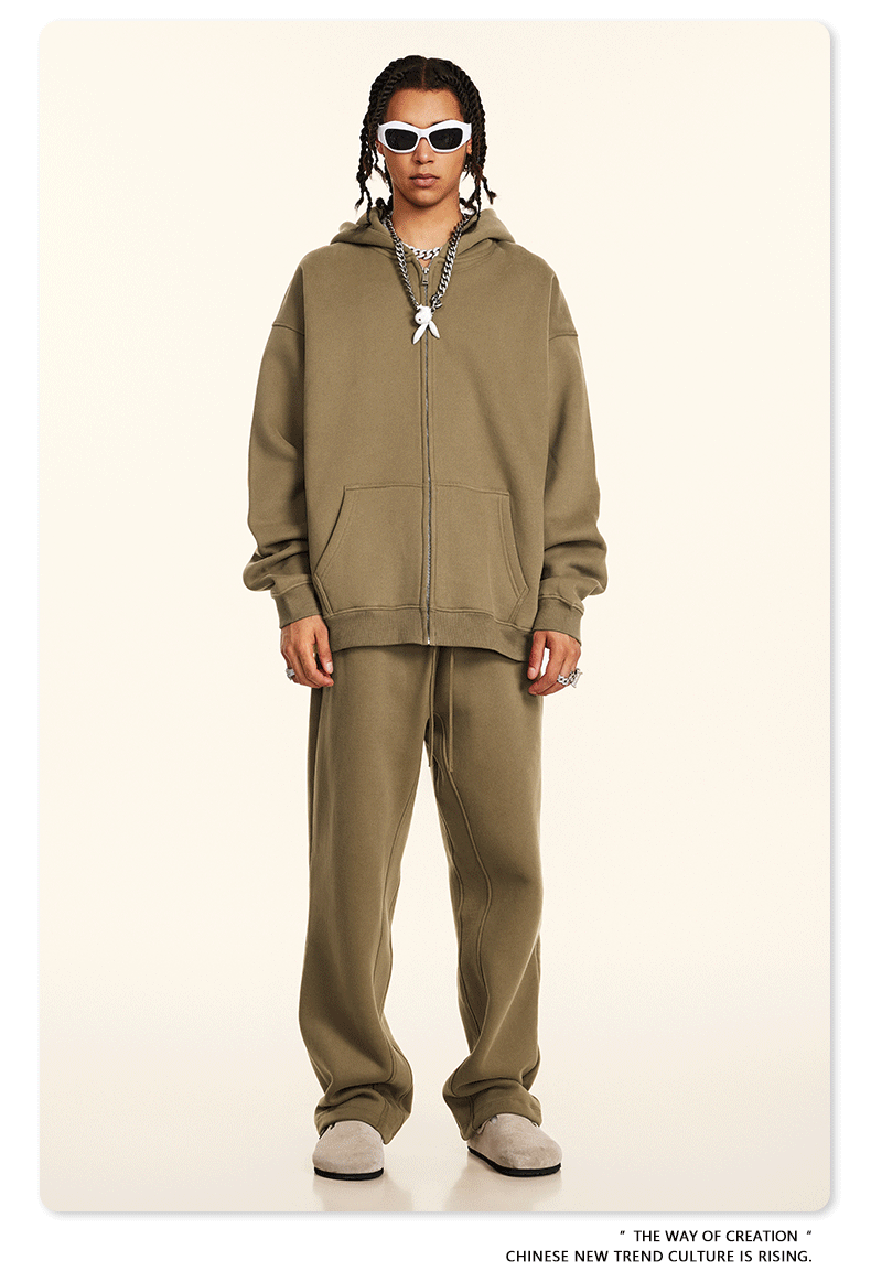 Model wearing olive tan oversized zip-up hoodie and matching relaxed sweatpants with white sunglasses, silver chains and beige shoes against cream backdrop