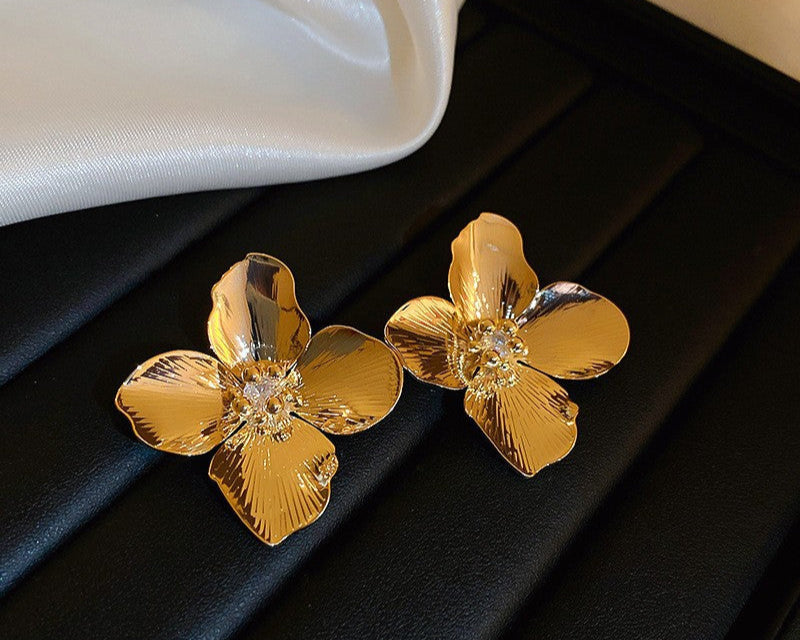 Glossy Gold Flower Earrings – Large gold floral earrings with reflective petals and a crystal center, placed on black fabric