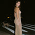 Woman in a shimmering nude gown standing on a city street at night, illuminated by car headlights