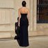 Back view of a woman wearing a strapless black evening gown with ruched details, standing in front of a textured stone wall