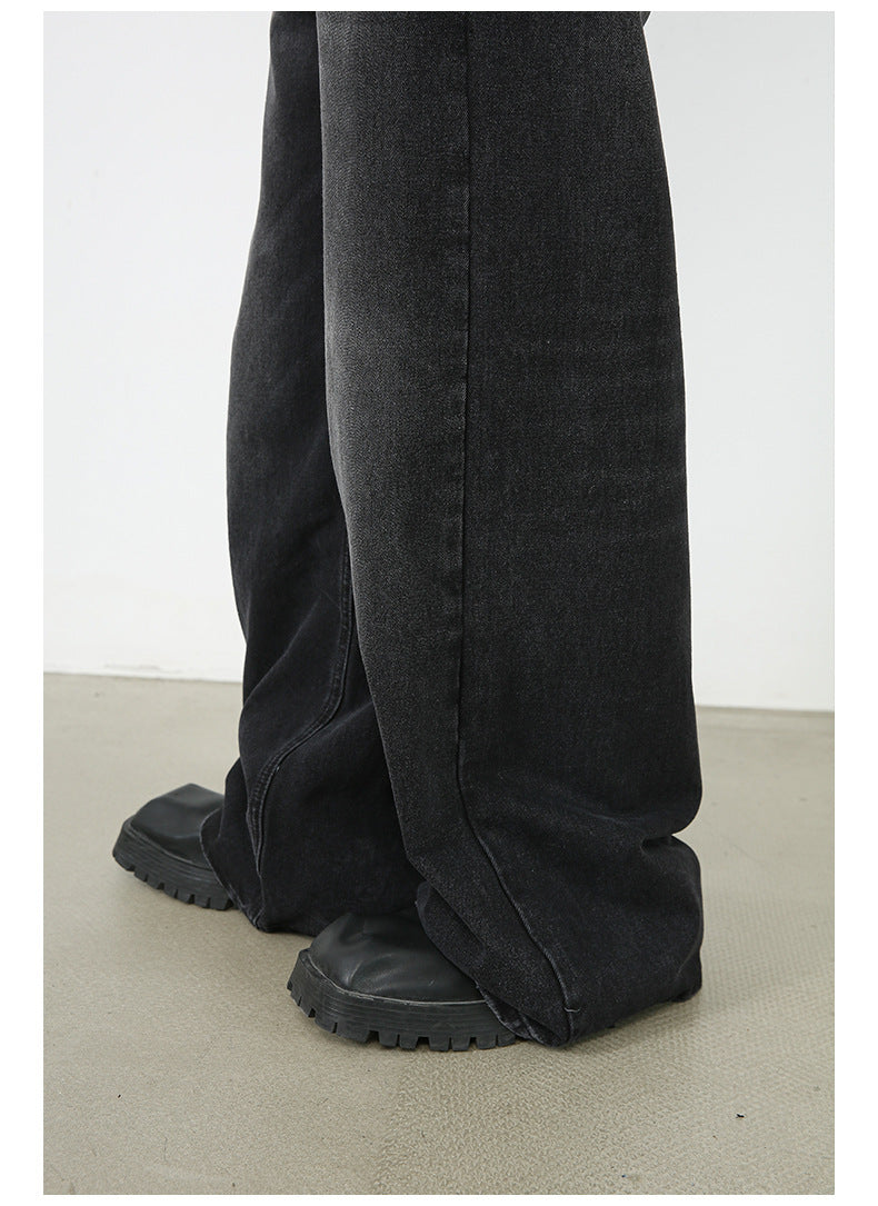 A close-up of black wide-leg denim jeans pooling at ankles over chunky platform shoes. Shows detailed hem construction and extra-long silhouette against neutral floor