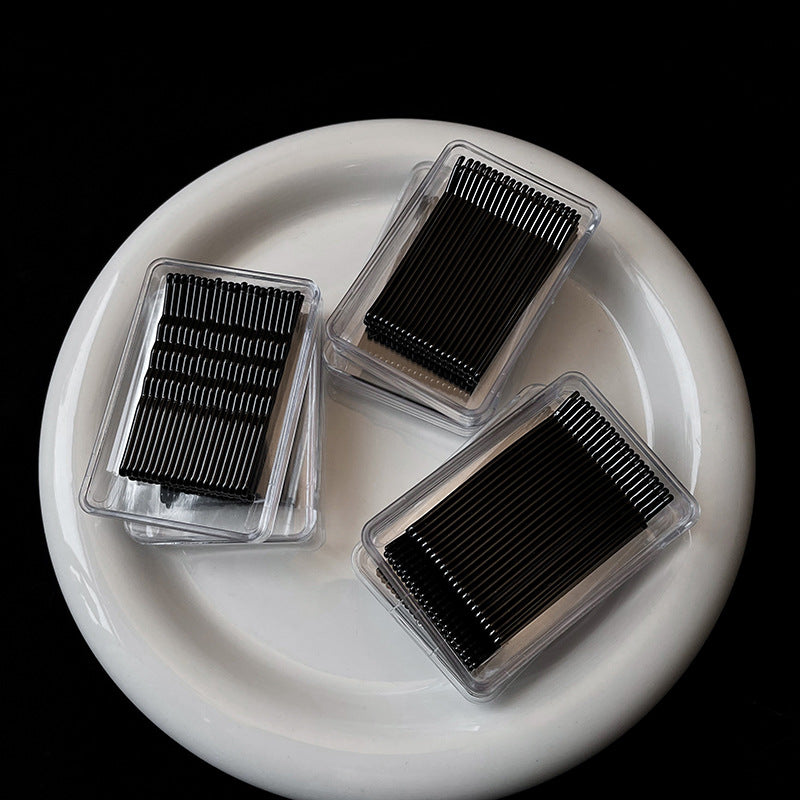 Three clear plastic boxes containing black bobby pins arranged on white ceramic plate, professional hair styling supplies