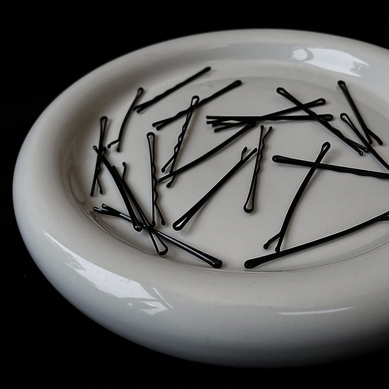 Black bobby pins scattered on white ceramic dish, minimalist hair accessories styling flatlay