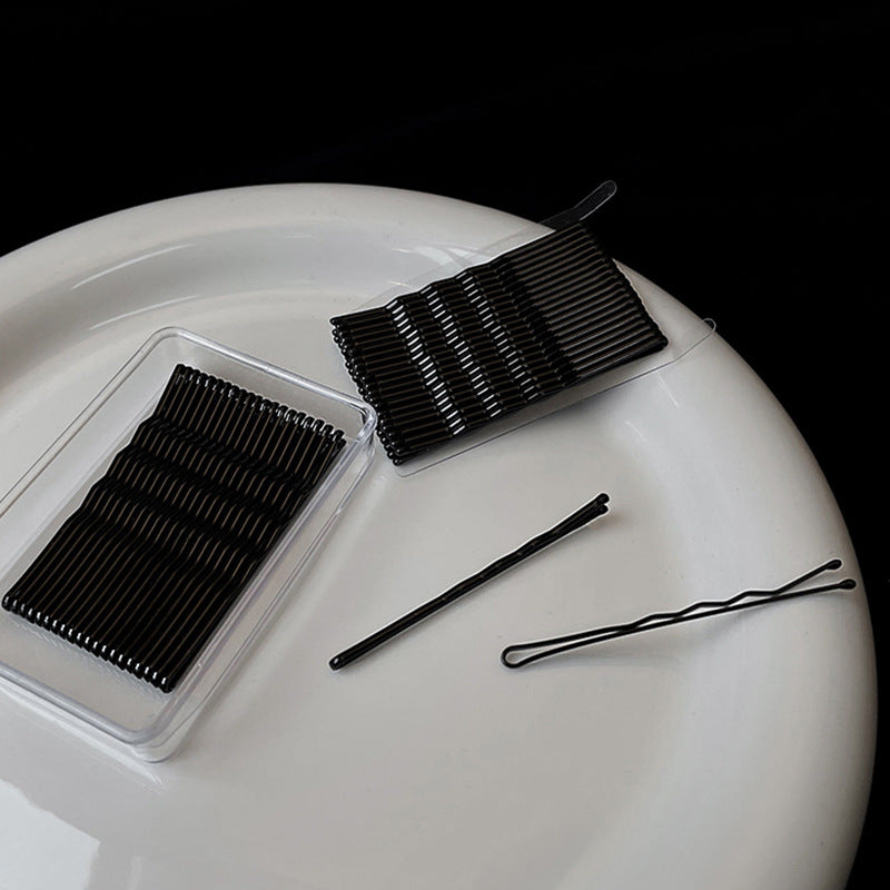 Two packs of black bobby pins with loose pins on white ceramic surface, minimalist beauty flatlay