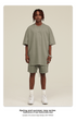 Model with dreadlocks wearing sage green t-shirt and matching shorts set with silver chain necklace and white sneakers against cream background