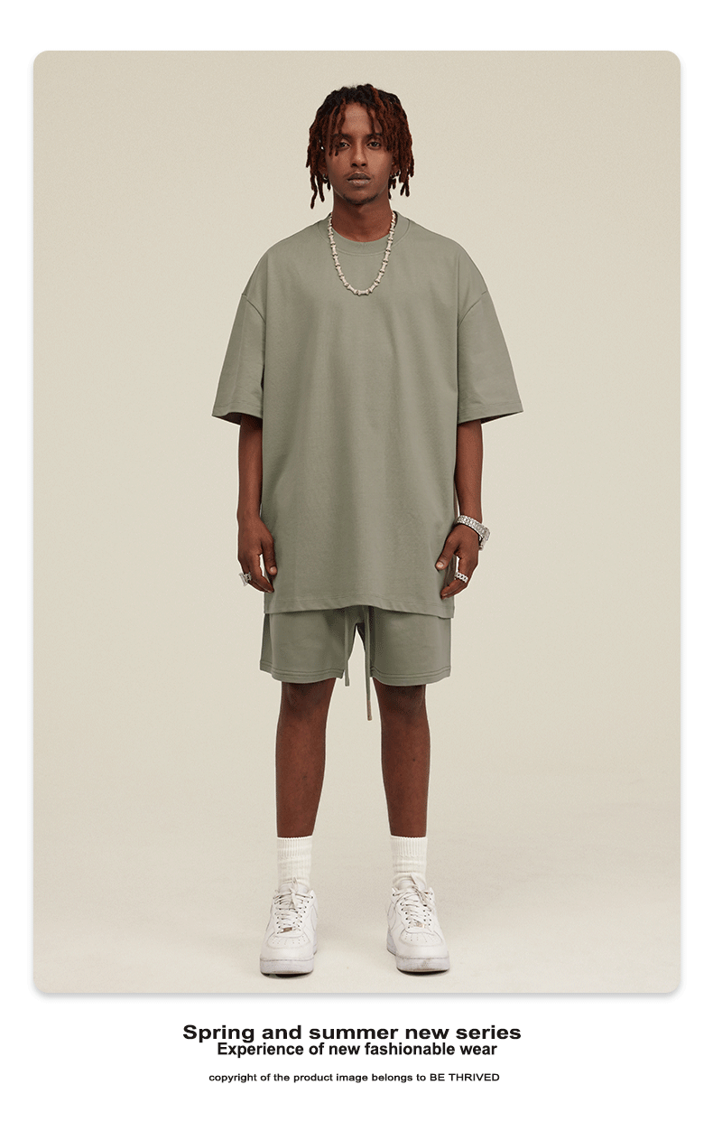 Model with dreadlocks wearing sage green t-shirt and matching shorts set with silver chain necklace and white sneakers against cream background
