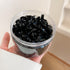 Hand holding clear container filled with small black plastic hair ties or elastics in a minimalist style