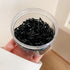 Hand holding clear plastic container filled with small black elastic hair ties, showing glossy texture and tangled arrangement