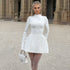 A woman in a white structured mini dress with flared skirt poses in front of stone columns, holding a quilted white handbag