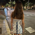 A model with long blonde hair is viewed from behind wearing a blue and white toile-print mini dress with corset back lacing, on a city street at night