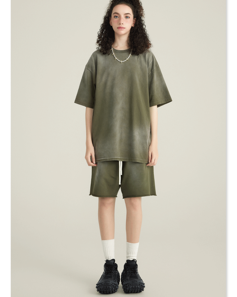 Model with curly hair wearing olive green t-shirt and matching shorts with white socks and black sneakers against cream background