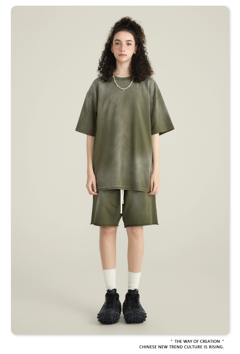 Model with curly hair wearing olive green t-shirt and matching shorts with white socks and black sneakers against cream background