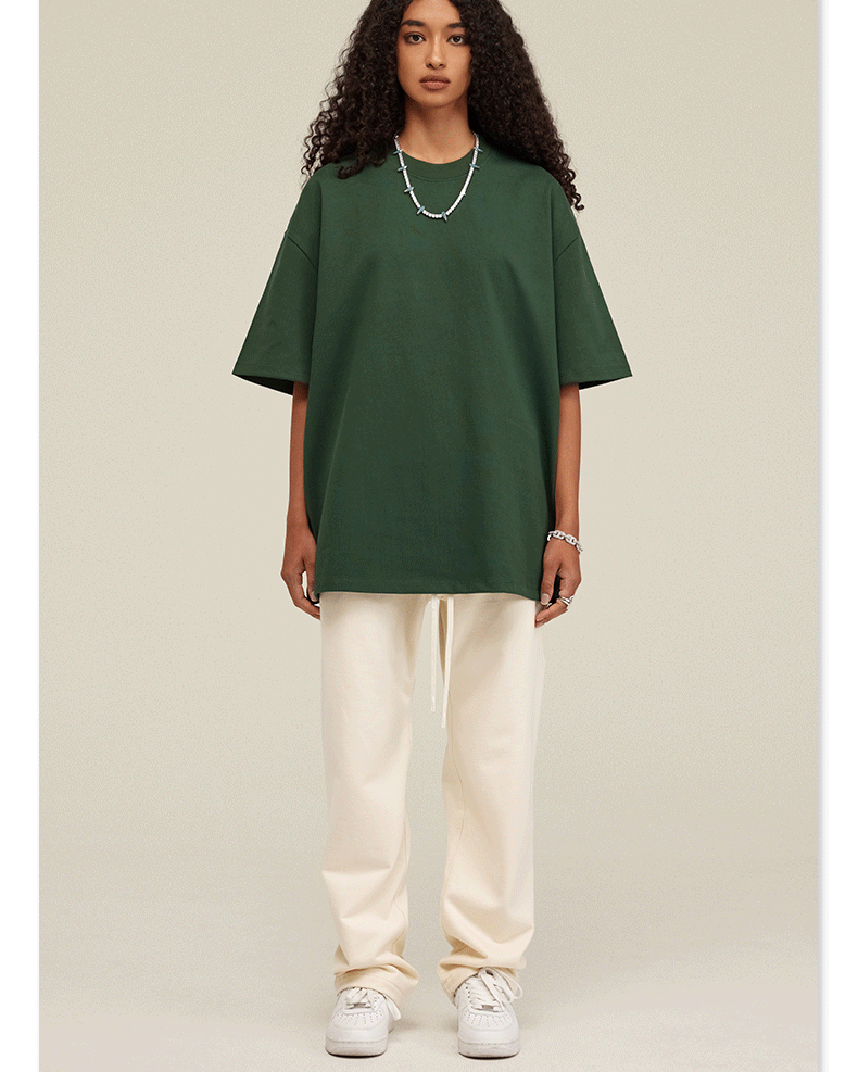 Woman wearing oversized dark green t-shirt with cream sweatpants, white sneakers and beaded necklace against neutral backdrop